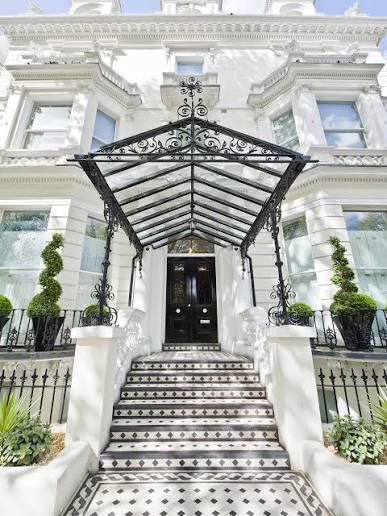 The entrance to a stucco house in Holland Park: a glazed ironwork canopy over a black panelled door, above chequered tiled steps between clipped topiary.