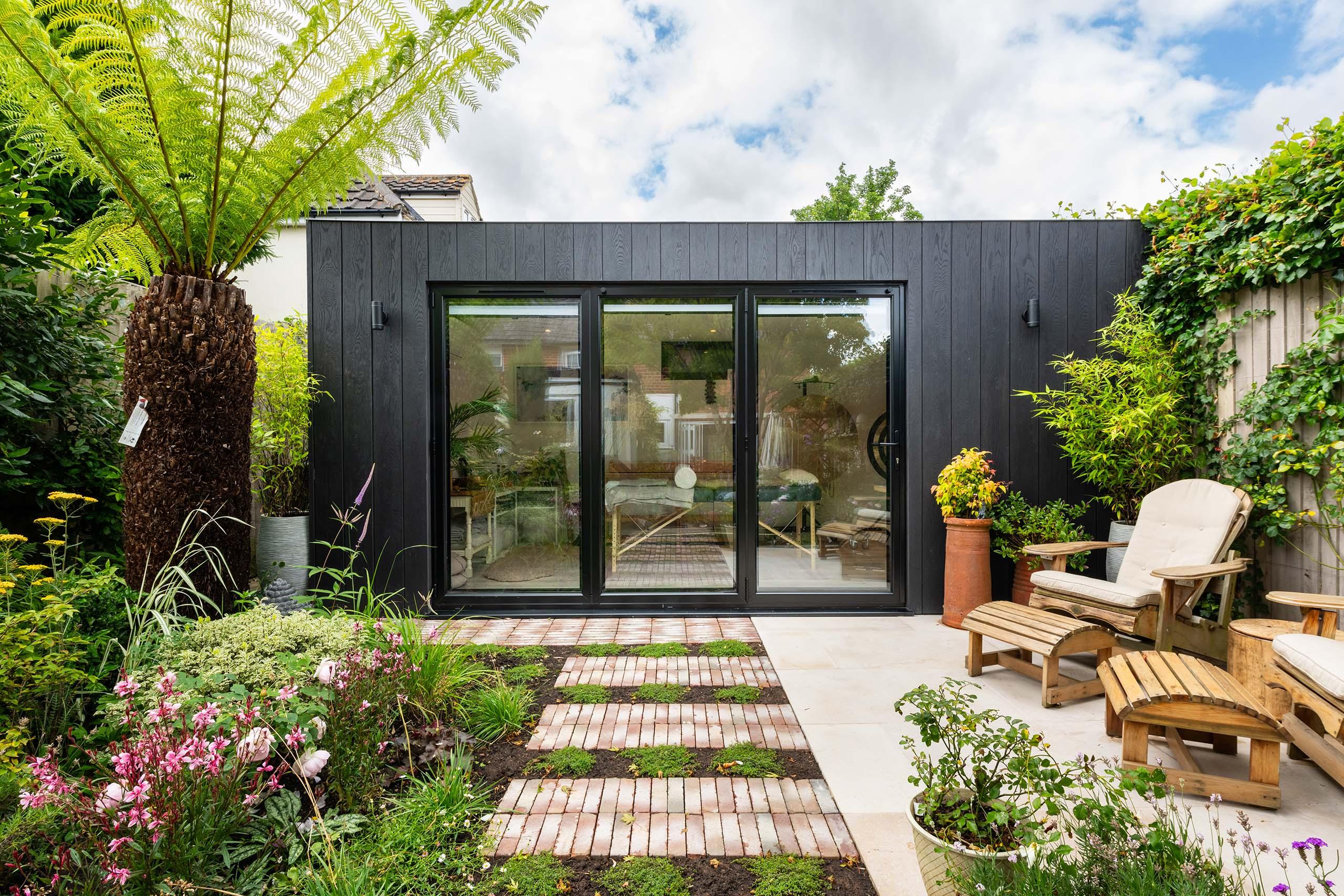 A black-clad garden building with full-height sliding glazing, set behind a brick and moss pathway.