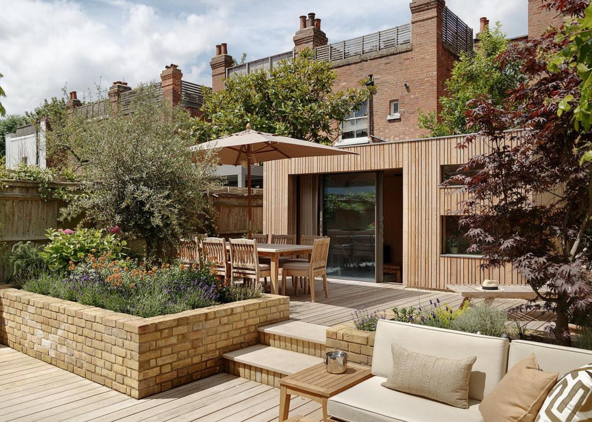 A timber-clad garden room opening onto a raised deck, with brick-edged planting beds and a mature acer.
