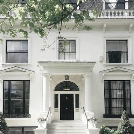 A white stuccoed frontage with a pedimented porch, black panelled door and clipped bay trees at the steps.